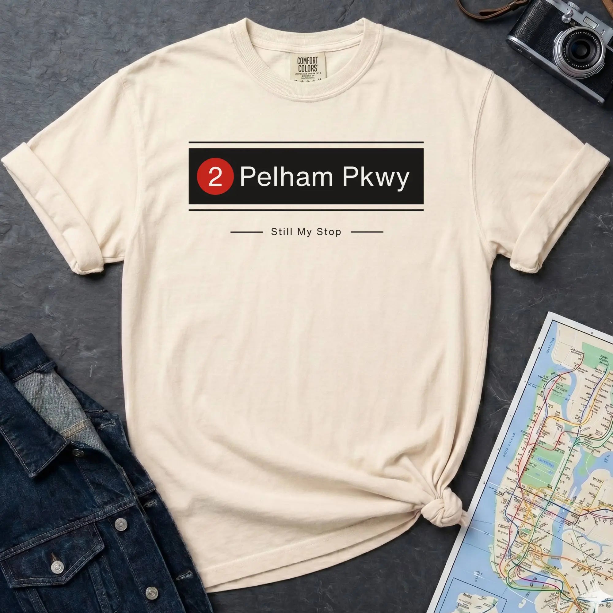 Ivory t-shirt with '2 Pelham Pkwy Still My Stop' design on a dark surface with denim jacket and map.