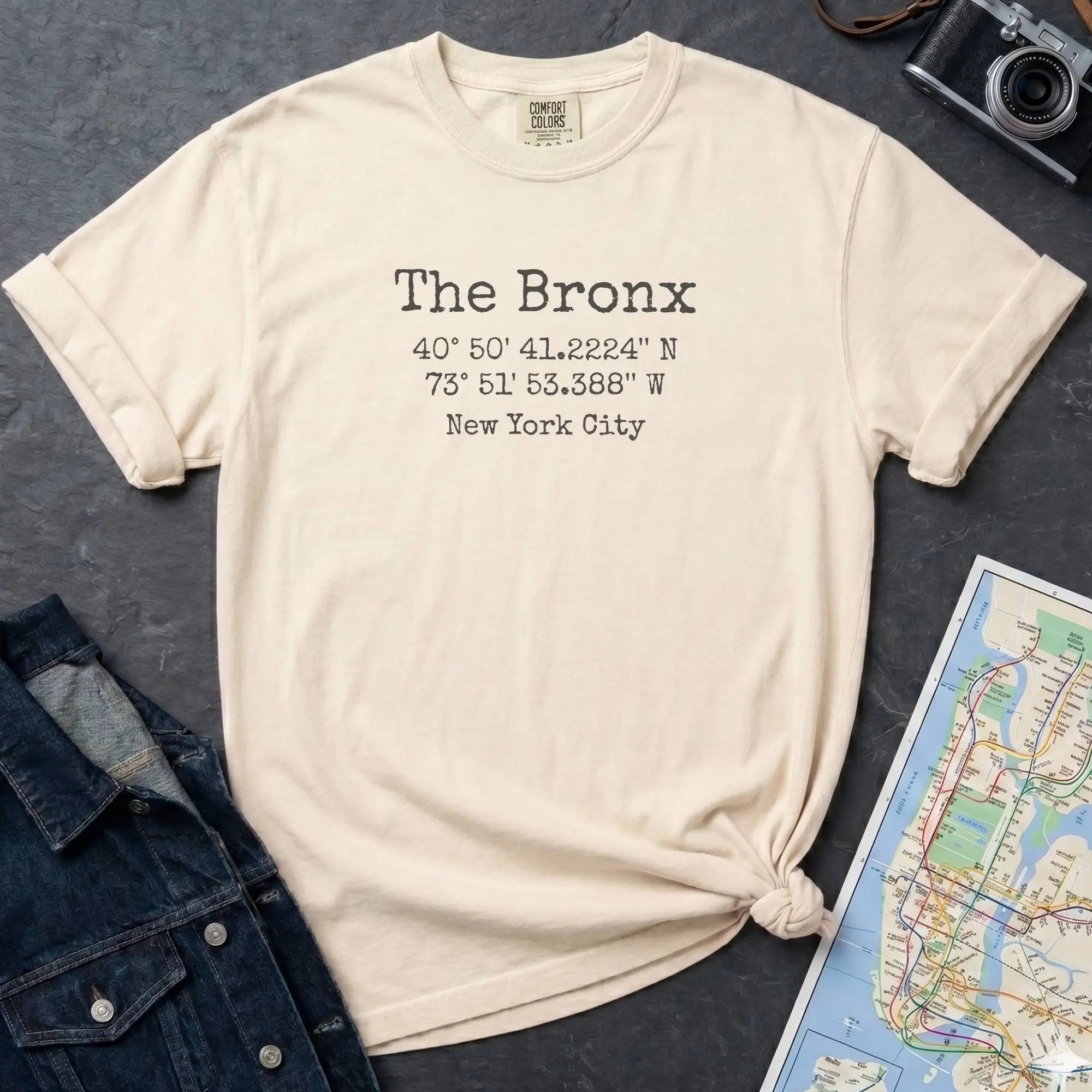 Beige t-shirt with 'The Bronx' text and coordinates, placed on a dark surface with a map and denim jacket.