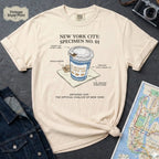 Ivory Comfort Colors t-shirt featuring a vintage New York City Anthora coffee cup graphic with specimen-style callouts and “The Official Chalice of New York” text.