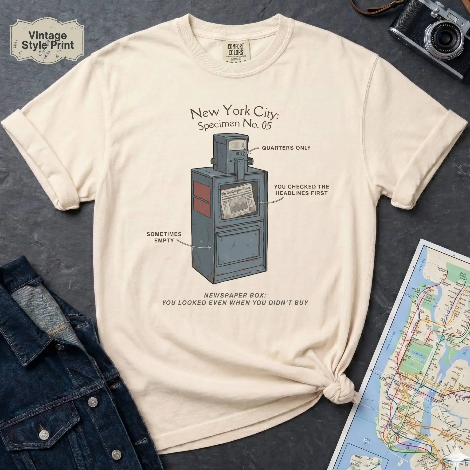 Ivory t-shirt with a graphic design of a vintage newspaper box and text on a gray surface with a map and camera.