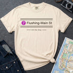 Beige t-shirt with 'Flushing-Main St' design on a dark surface with a denim jacket and map.