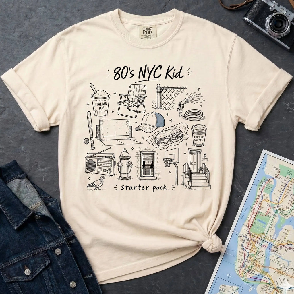 Ivory Comfort Colors t-shirt featuring an 80’s NYC Kid Starter Pack graphic with hand-drawn New York City icons including Italian ice, a radio, stoop steps, stickball, a fire hydrant, and a pigeon.