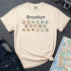 Ivory t-shirt with 'Brooklyn' and subway line numbers printed on it, placed on a dark surface with a map and denim jacket.