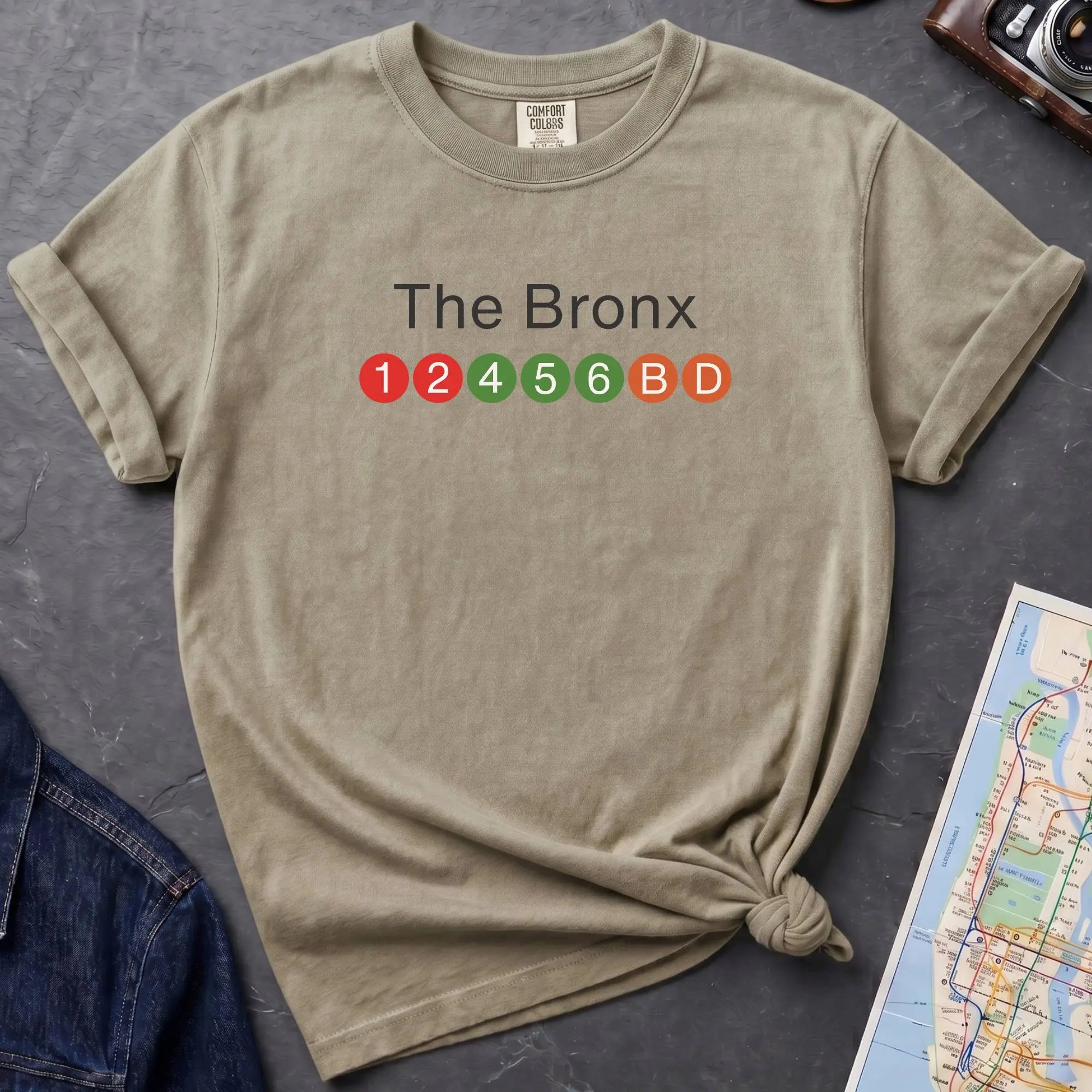 Khaki colored  t-shirt with 'The Bronx' text and NYC subway lines on a dark surface with a denim jacket, camera and a map.