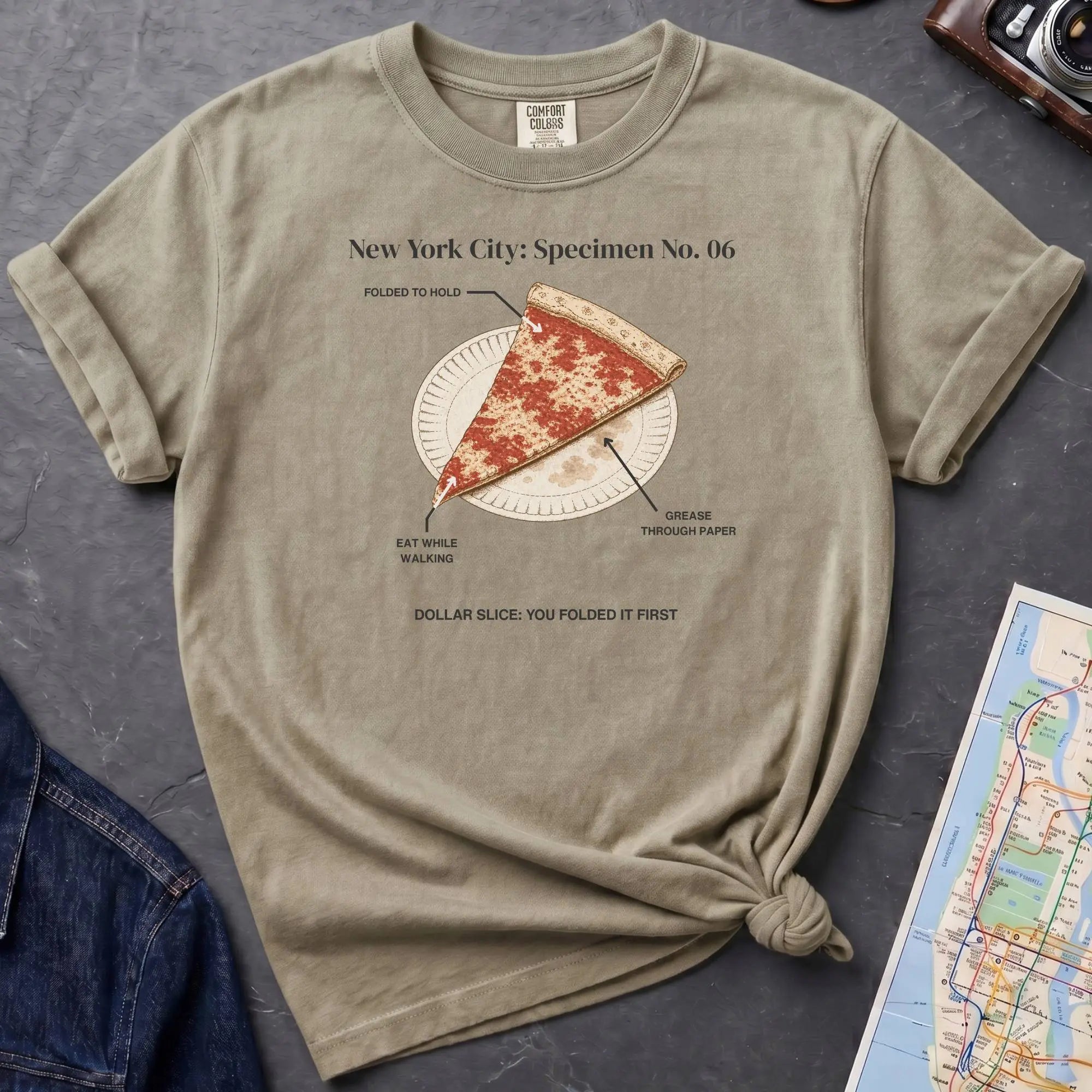 Khaki colored Comfort Colors t-shirt featuring a vintage New York City dollar slice pizza graphic with specimen-style callouts and a folded slice on a paper plate.