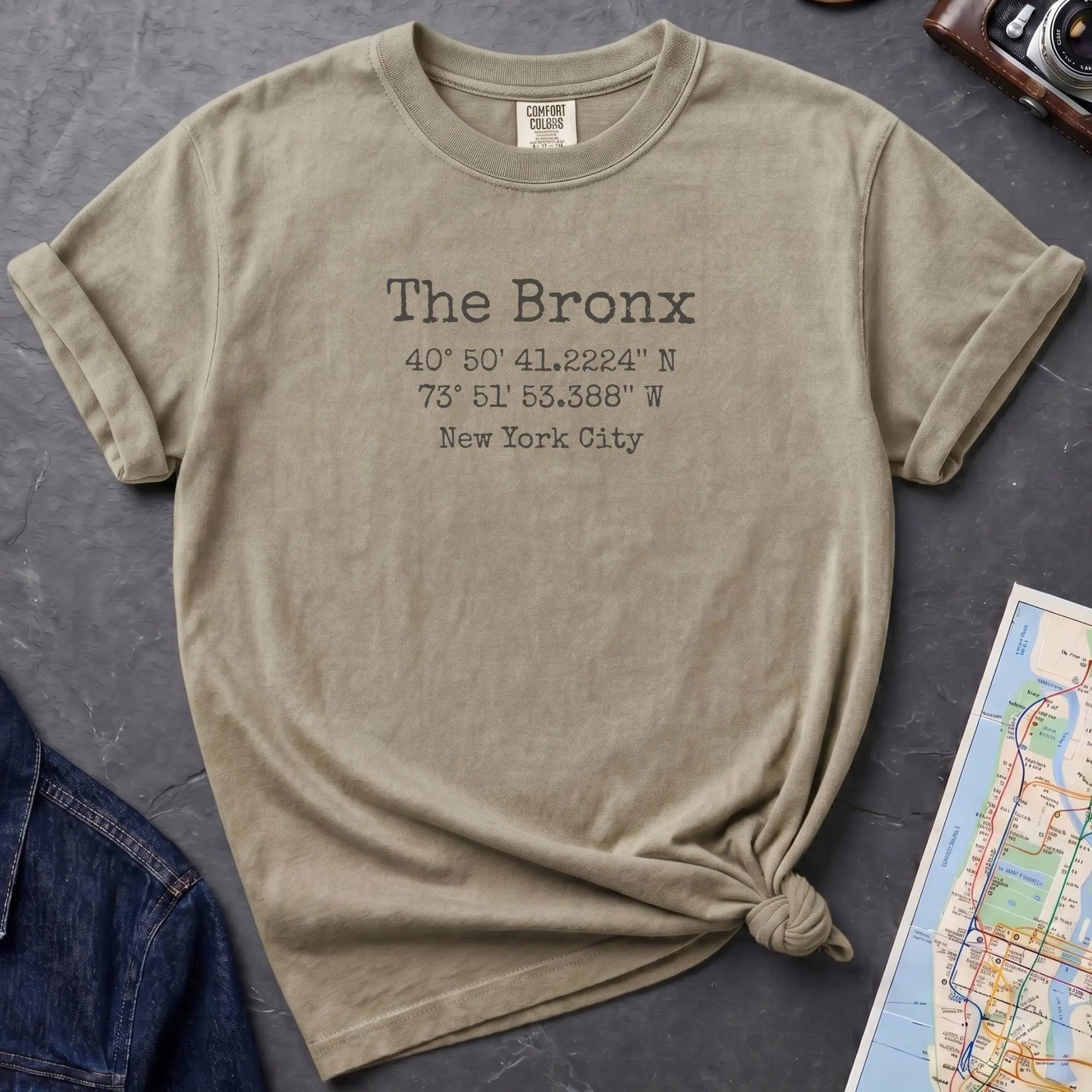 Khaki colored t-shirt with 'The Bronx' text and coordinates, placed on a dark surface with a map and denim jacket.