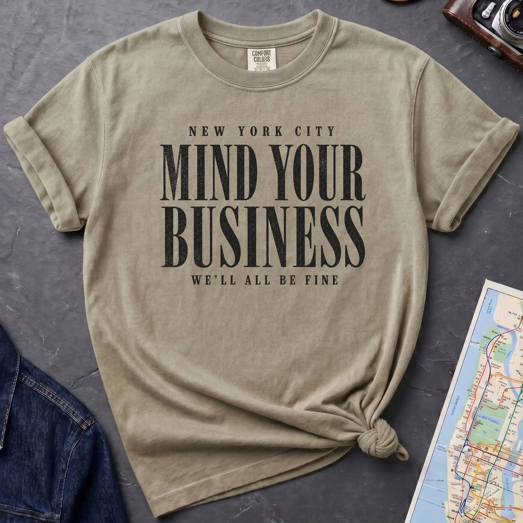 Khaki colored Comfort Colors t-shirt featuring bold New York City “Mind Your Business, We’ll All Be Fine” text in a vintage black typography design.