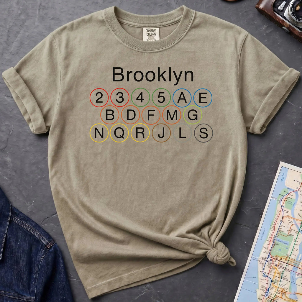 Khaki colored t-shirt with 'Brooklyn' and subway line numbers printed on it, placed on a dark surface with a map and denim jacket.