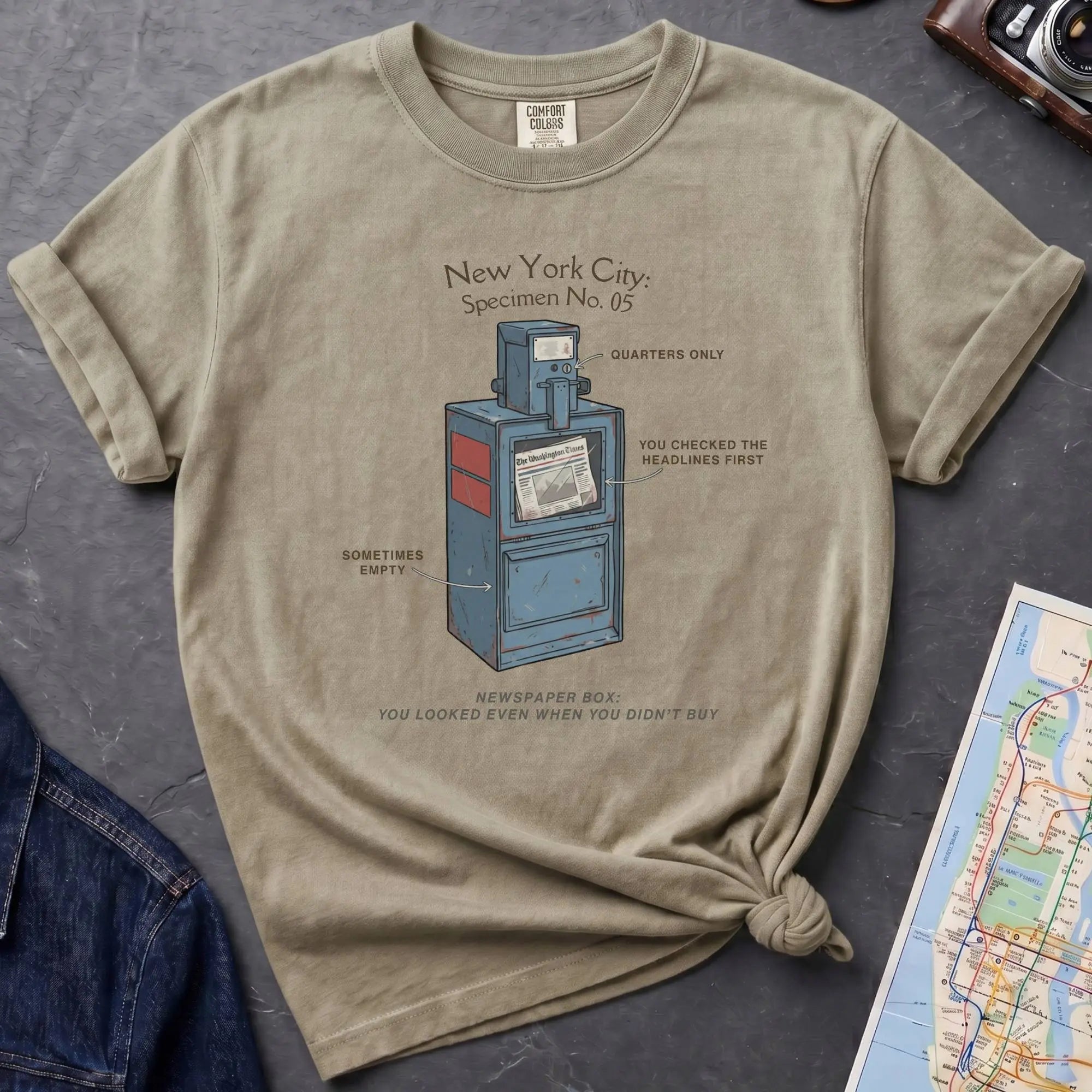 Khaki colored t-shirt with a newspaper box graphic and text on a textured surface with a camera and map.