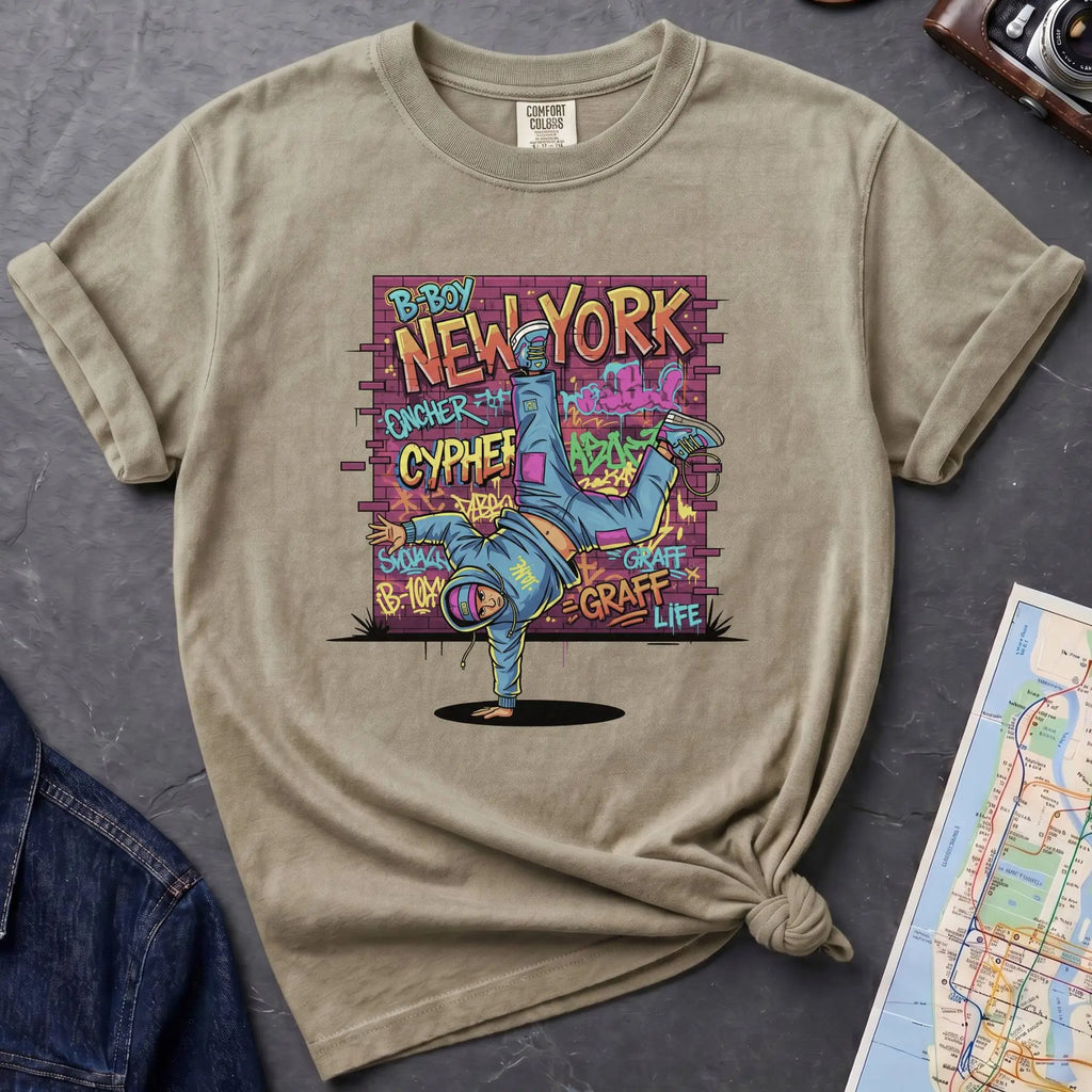 Khaki colored t-shirt with a colorful graphic design featuring a breakdancer and text on a dark surface with a denim jacket and a map.