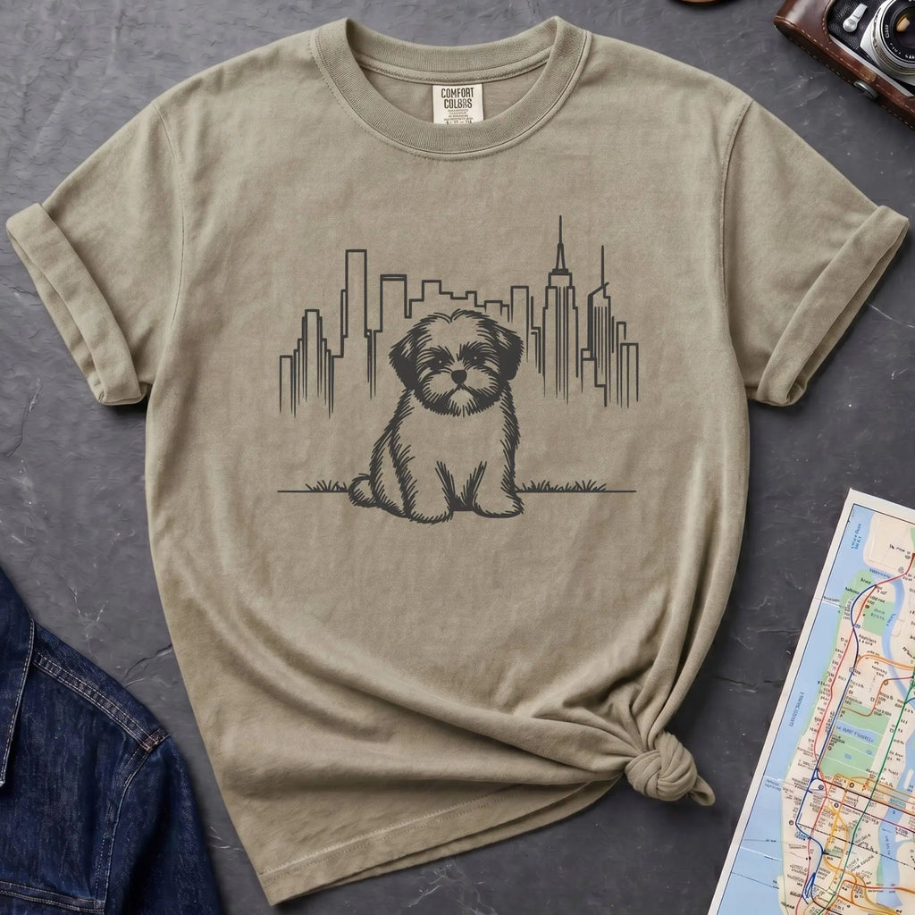 Khaki colored t-shirt with a Shih Tzu dog and cityscape design on a dark surface with a map and camera.