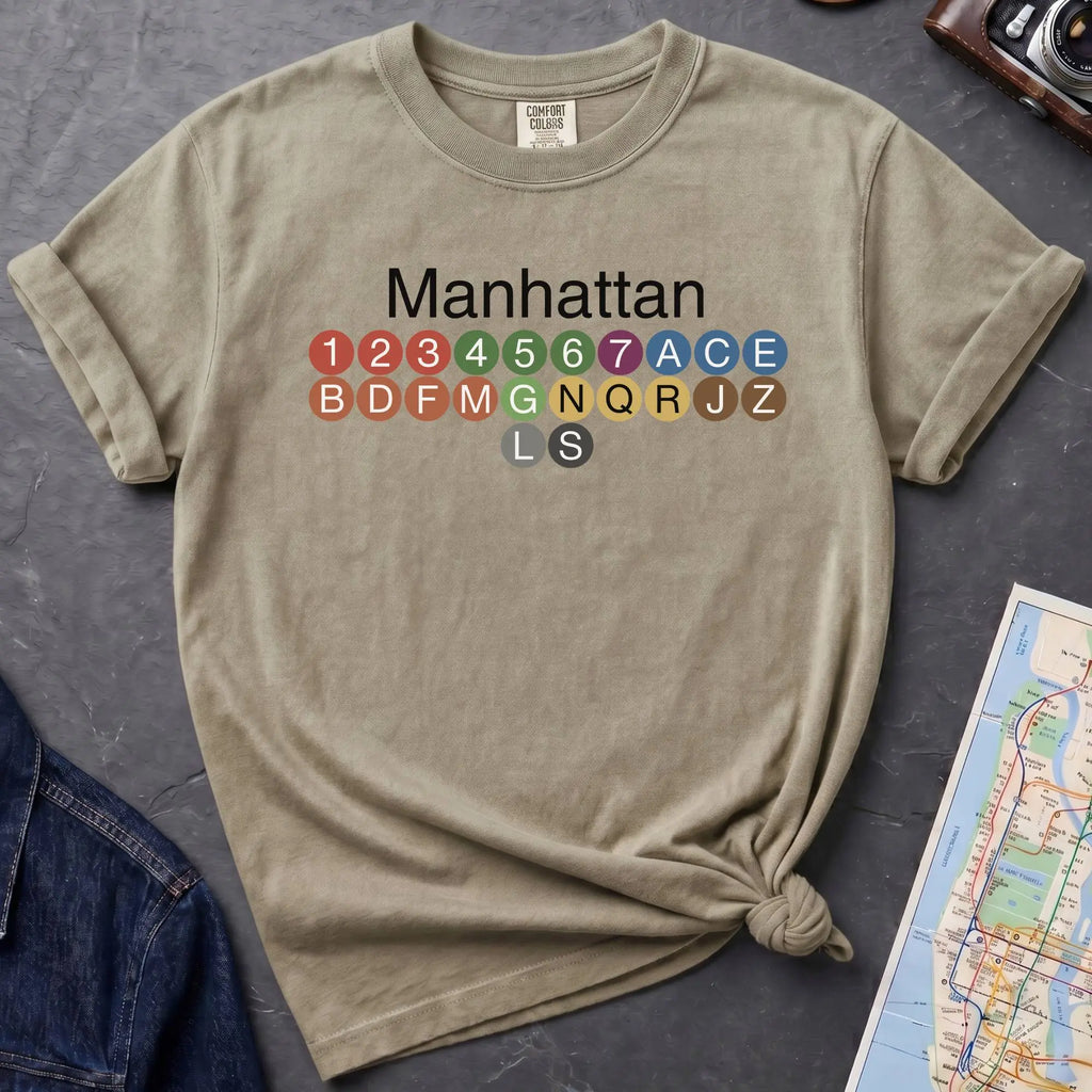 Khaki colored Comfort Colors t-shirt featuring a minimalist Manhattan subway line graphic with colorful NYC train icons.