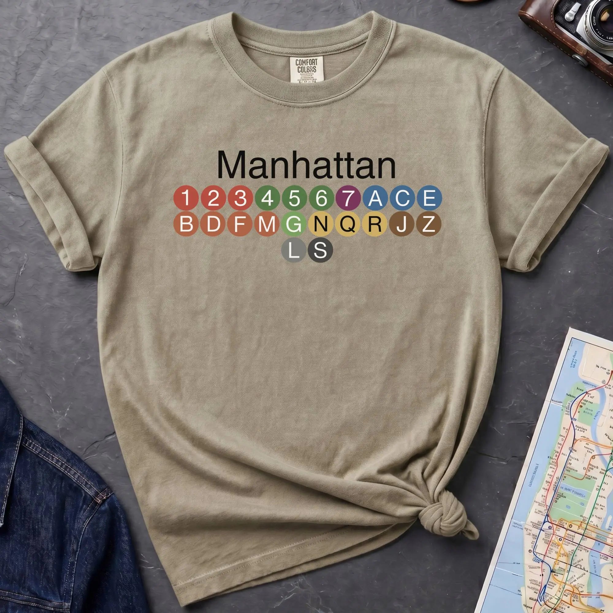 Khaki colored Comfort Colors t-shirt featuring a minimalist Manhattan subway line graphic with colorful NYC train icons.