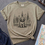 Khaki colored t-shirt with a Golden Doodle dog and cityscape design on a gray surface with a map and camera.