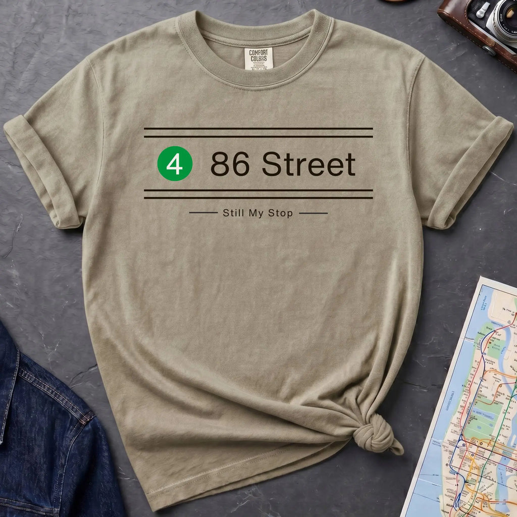 Beige t-shirt with '86 Street Still My Stop' design on a dark surface with a map and jacket.