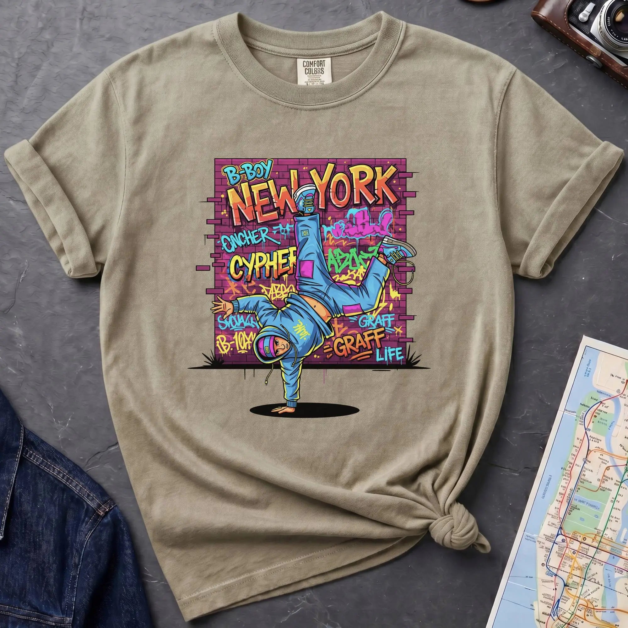 Khaki colored t-shirt with a colorful graphic design featuring a breakdancer and text on a dark surface with a denim jacket and a map.