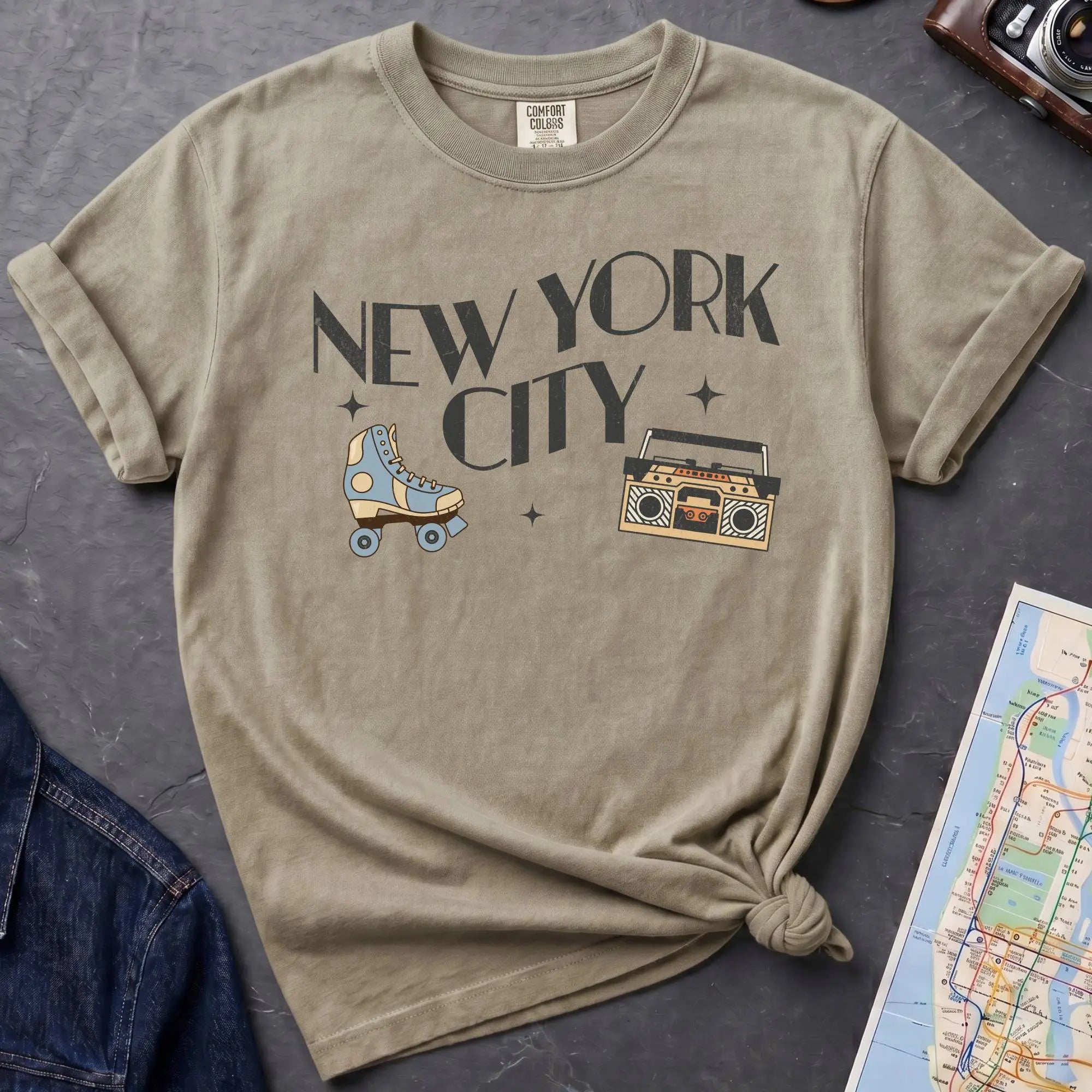 Khaki colored t-shirt with 'New York City' text and a roller skate and boom box on a dark surface with a map and camera.