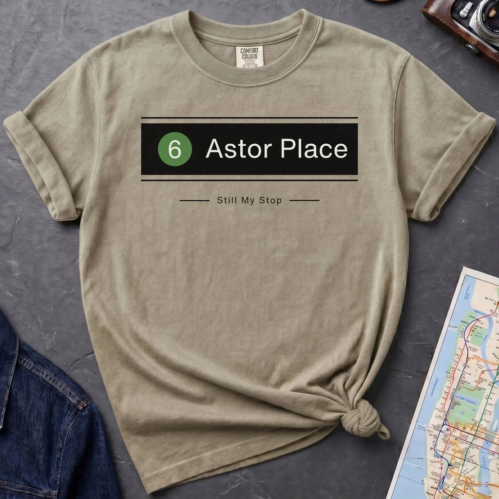Khaki colored t-shirt with '6 Astor Place Still My Stop' design on a dark surface with a map and a denim jacket.