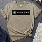 Khaki colored t-shirt with '6 Astor Place Still My Stop' design on a dark surface with a map and a denim jacket.