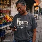 A woman wearing a pepper-colored Comfort Colors graphic t-shirt with 'Korvettes Discount Department Store New York City, Est. 1980' printed on the front, standing in an outdoor street setting and looking at her phone.