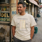 Man wearing Comfort Colors T-shirt with NYC library loan card specimen front print, while standing outside a deli on a city street
