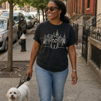 Woman walking a dog on a city street wearing a black Comfort Colors t-shirt with a Maltese dog and cityscape design.