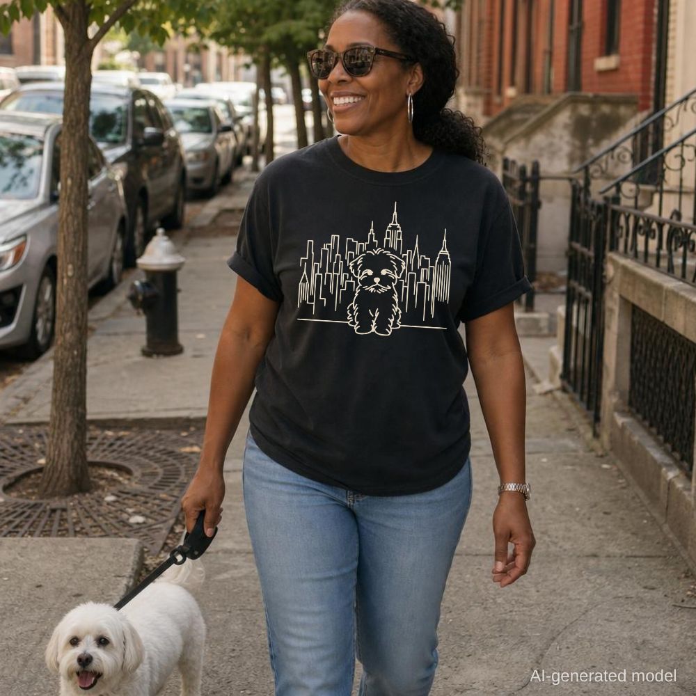 Woman walking a dog on a city street wearing a black Comfort Colors t-shirt with a Maltese dog and cityscape design.