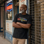 A man wearing a Black Comfort Colors t-shirt featuring a minimalist Manhattan subway line graphic with colorful NYC train icons while leaning against a storefront gate.