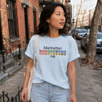 A woman wearing a Light blue Comfort Colors t-shirt featuring a minimalist Manhattan subway line graphic with colorful NYC train icons while walking down a street
