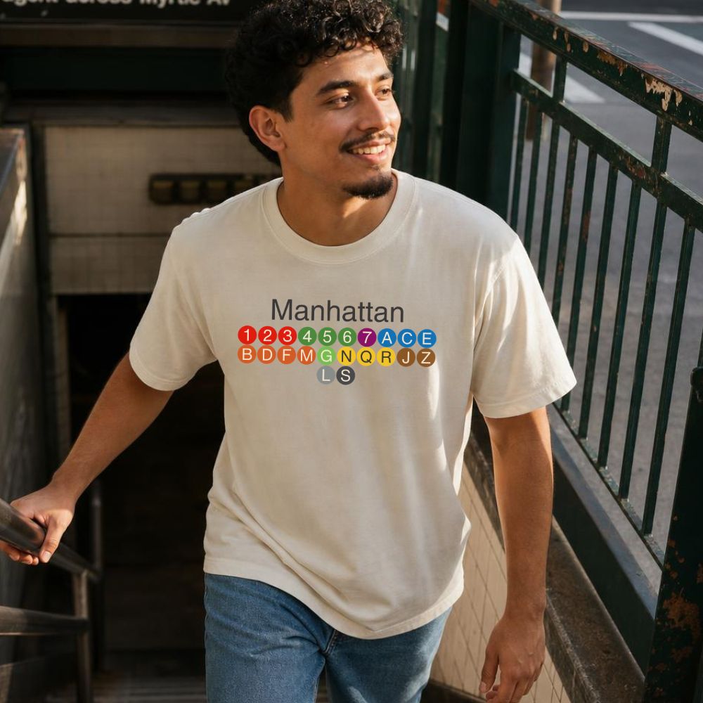 Man wearing an Ivory Comfort Colors t-shirt with Manhattan text and subway line design, standing outdoors.