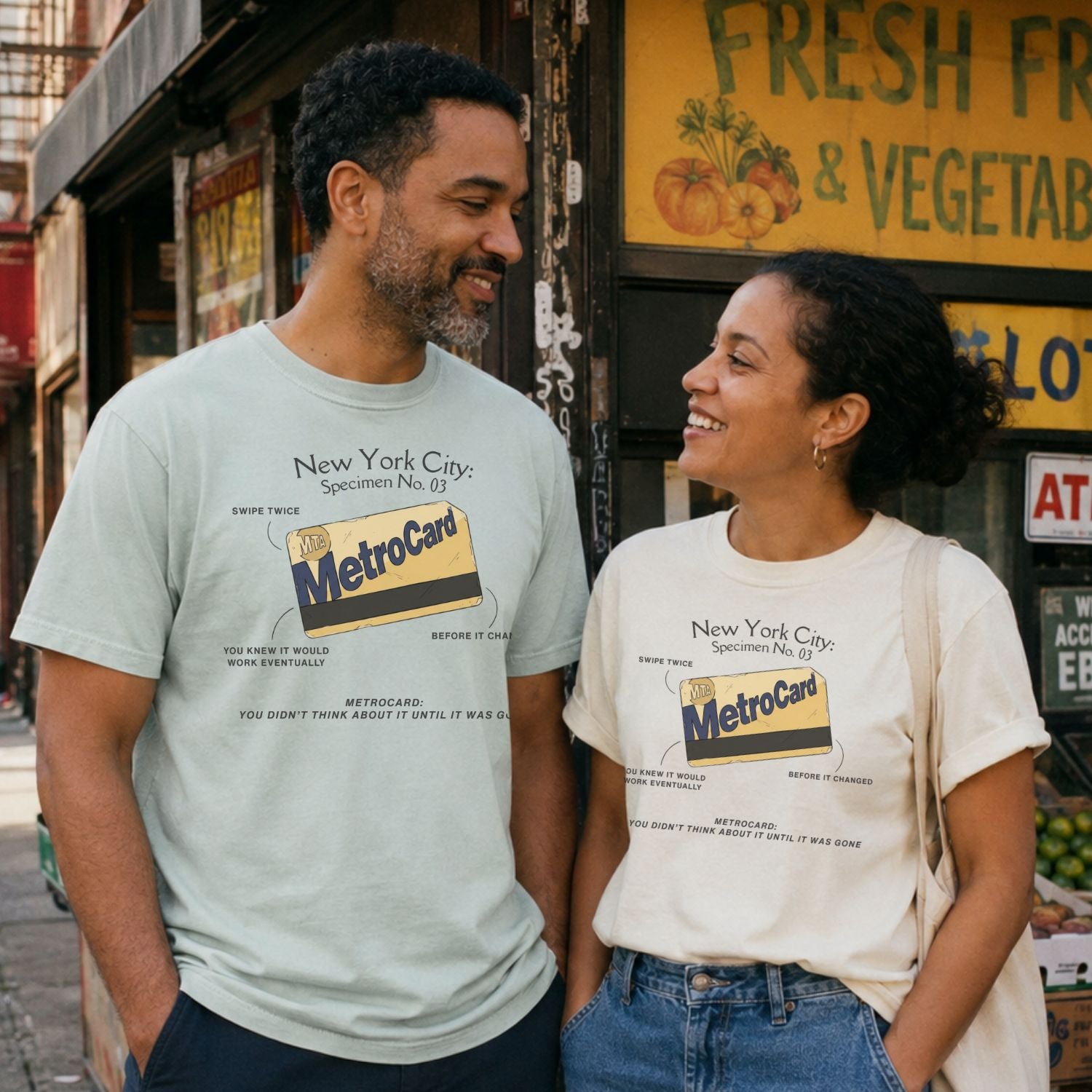 Two people wearing a Bay and an Ivory colored Comfort Colors t-shirt featuring a vintage New York City MetroCard graphic with specimen-style callouts and “You didn’t think about it until it was gone” text standing in front of a store.