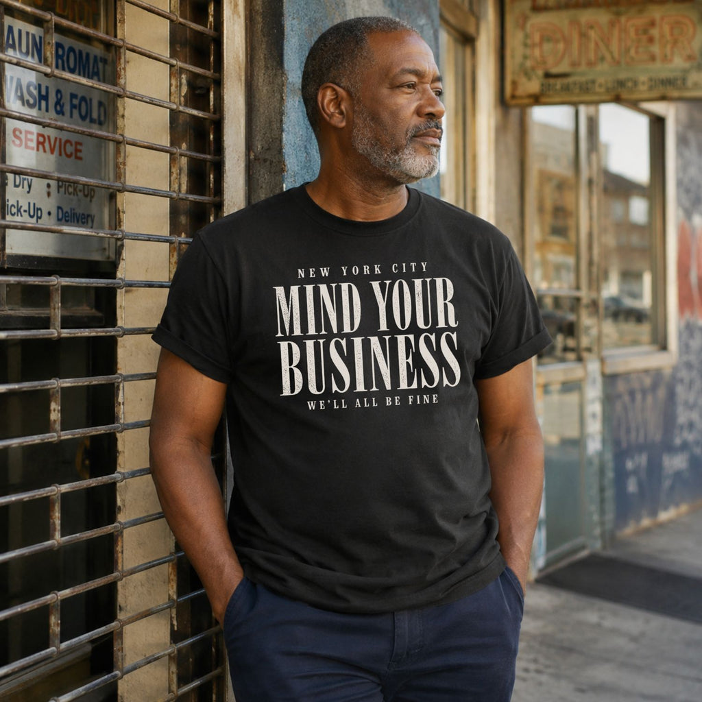 Man wearing a black t-shirt with 'New York City Mind Your Business We'll All Be Fine' text, standing against a building.