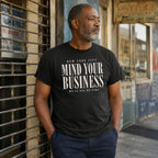 Man wearing a black t-shirt with 'New York City Mind Your Business We'll All Be Fine' text, standing against a building.