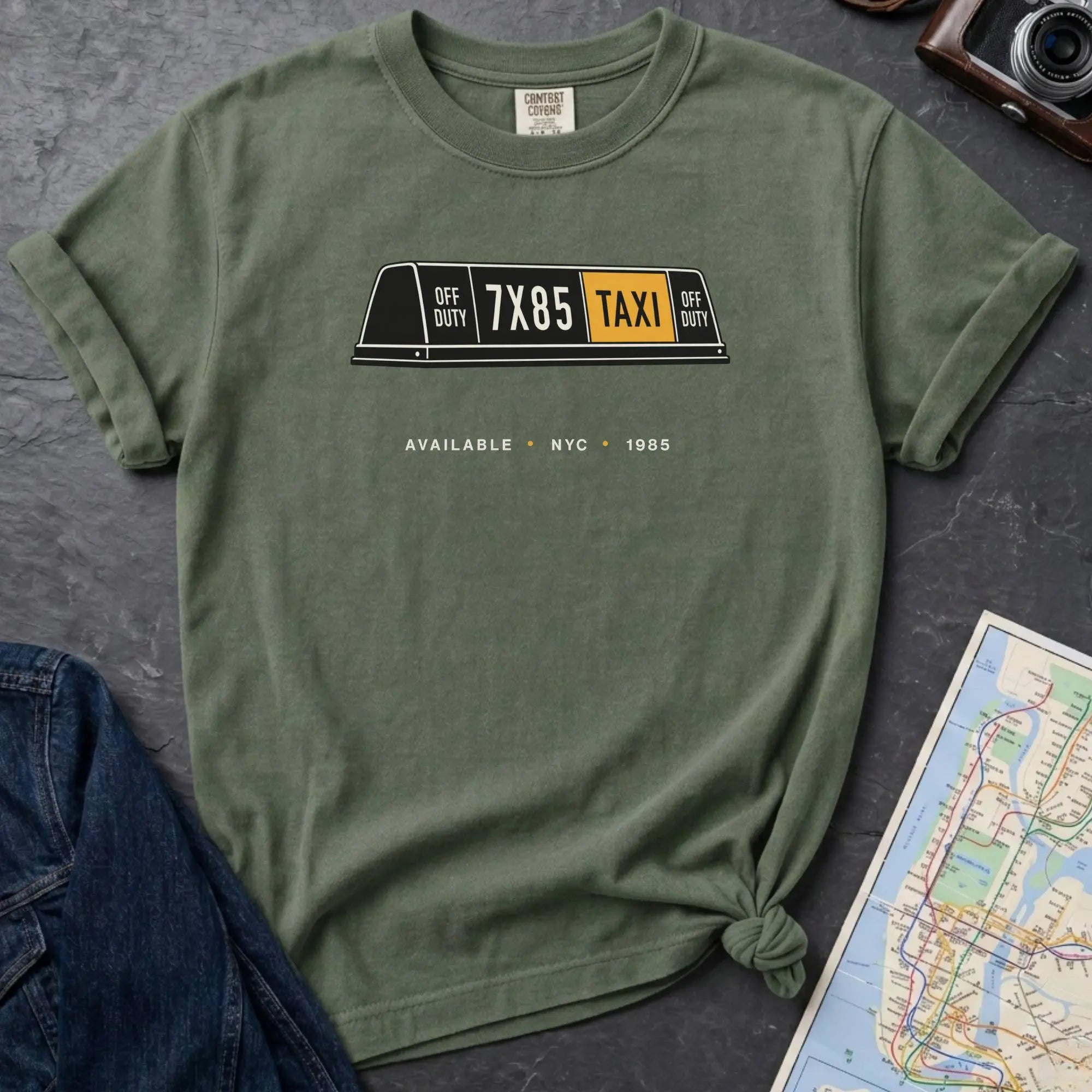 Moss green Comfort Colors t-shirt featuring a vintage New York City taxi roof sign graphic with “Available • NYC • 1985” text.