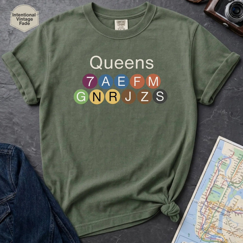 Moss green Black Comfort Colors t-shirt featuring a minimalist Queens subway line graphic with colorful NYC train icons.