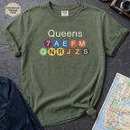 Moss green Black Comfort Colors t-shirt featuring a minimalist Queens subway line graphic with colorful NYC train icons.