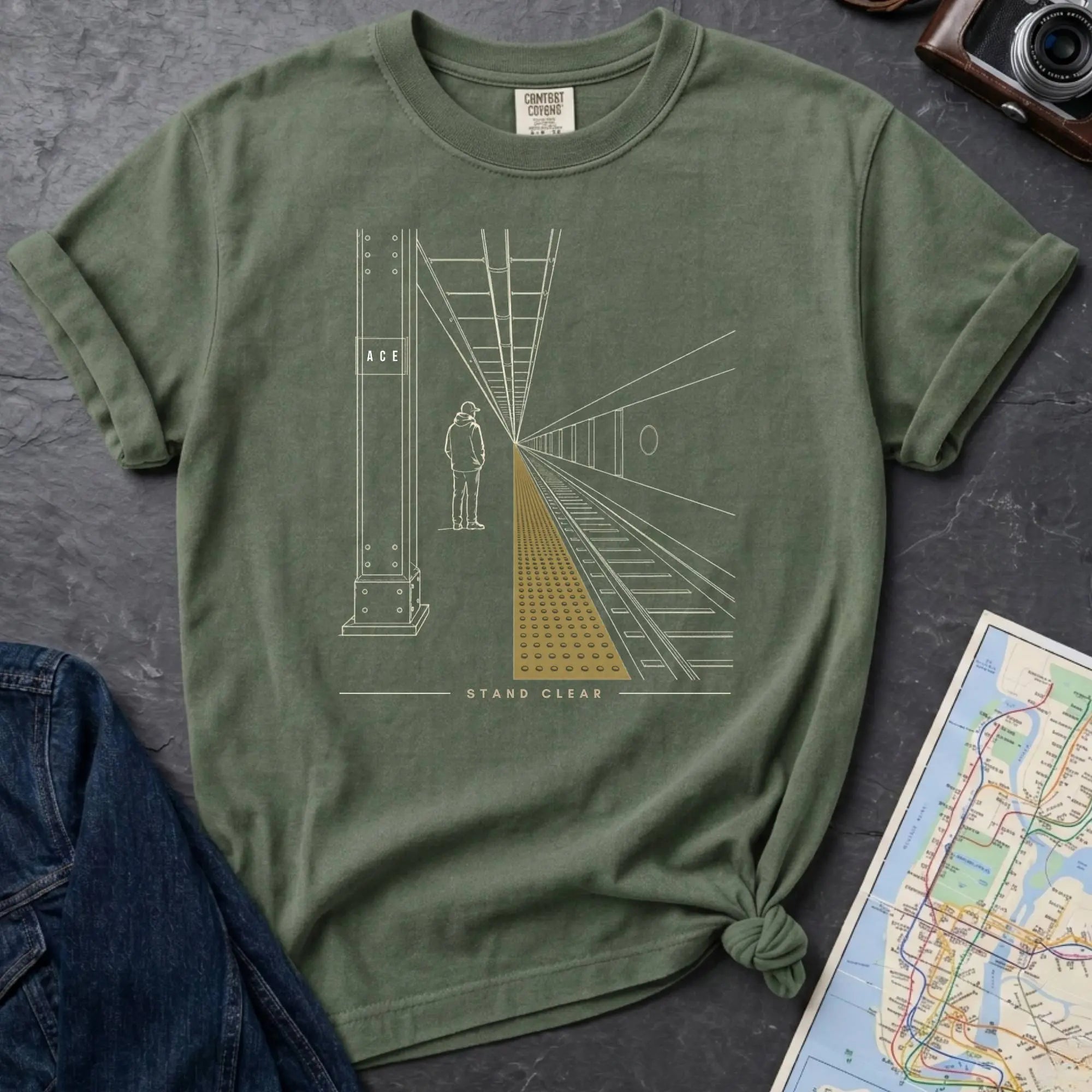 Moss green Comfort Colors t-shirt featuring a minimalist NYC subway platform graphic with a lone figure, train tracks, and “Stand Clear” text.