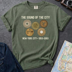 Moss green Comfort Colors t-shirt featuring vintage New York City subway tokens with “The Sound of the City” and “New York City • 1953–2003” text.