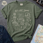 Moss Green t-shirt with '80s NYC Subway Survival Kit' graphic on a dark surface with a map and jeans.
