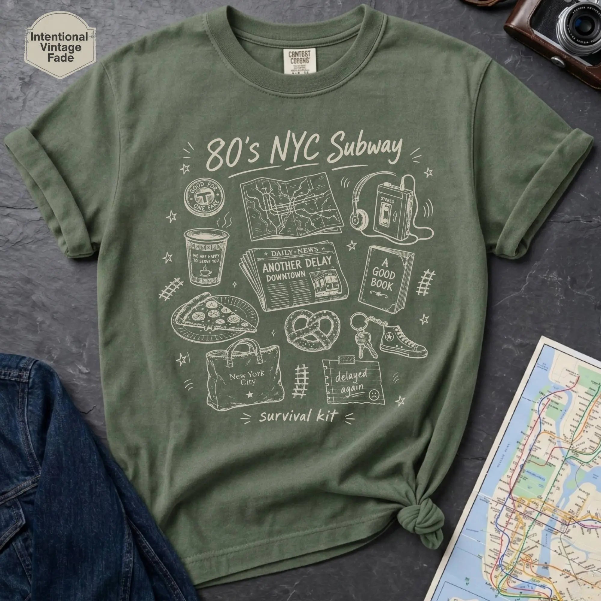 Moss Green t-shirt with '80s NYC Subway Survival Kit' graphic on a dark surface with a map and jeans.