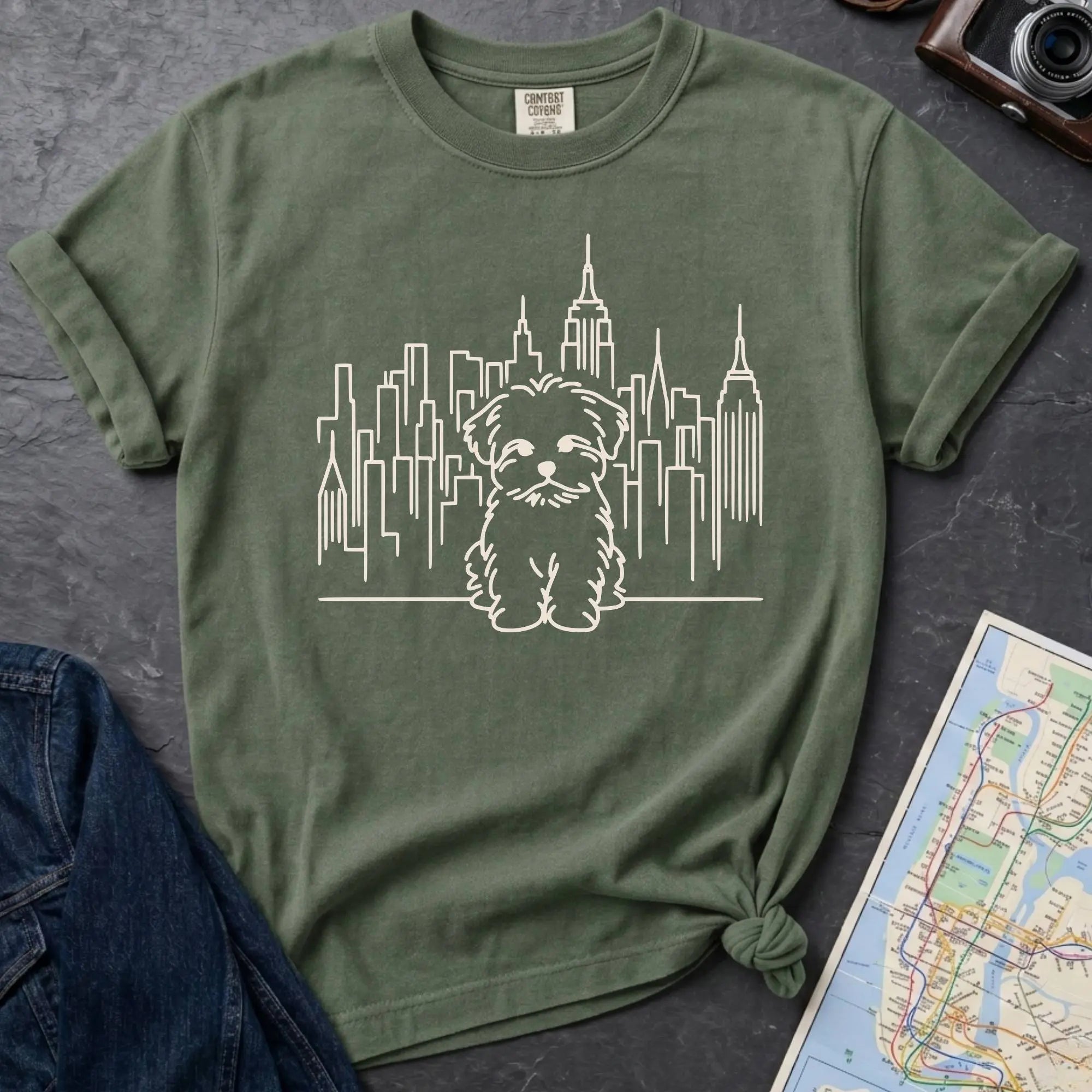 Moss green t-shirt with a Maltese dog and cityscape design on a gray surface with a map and camera.