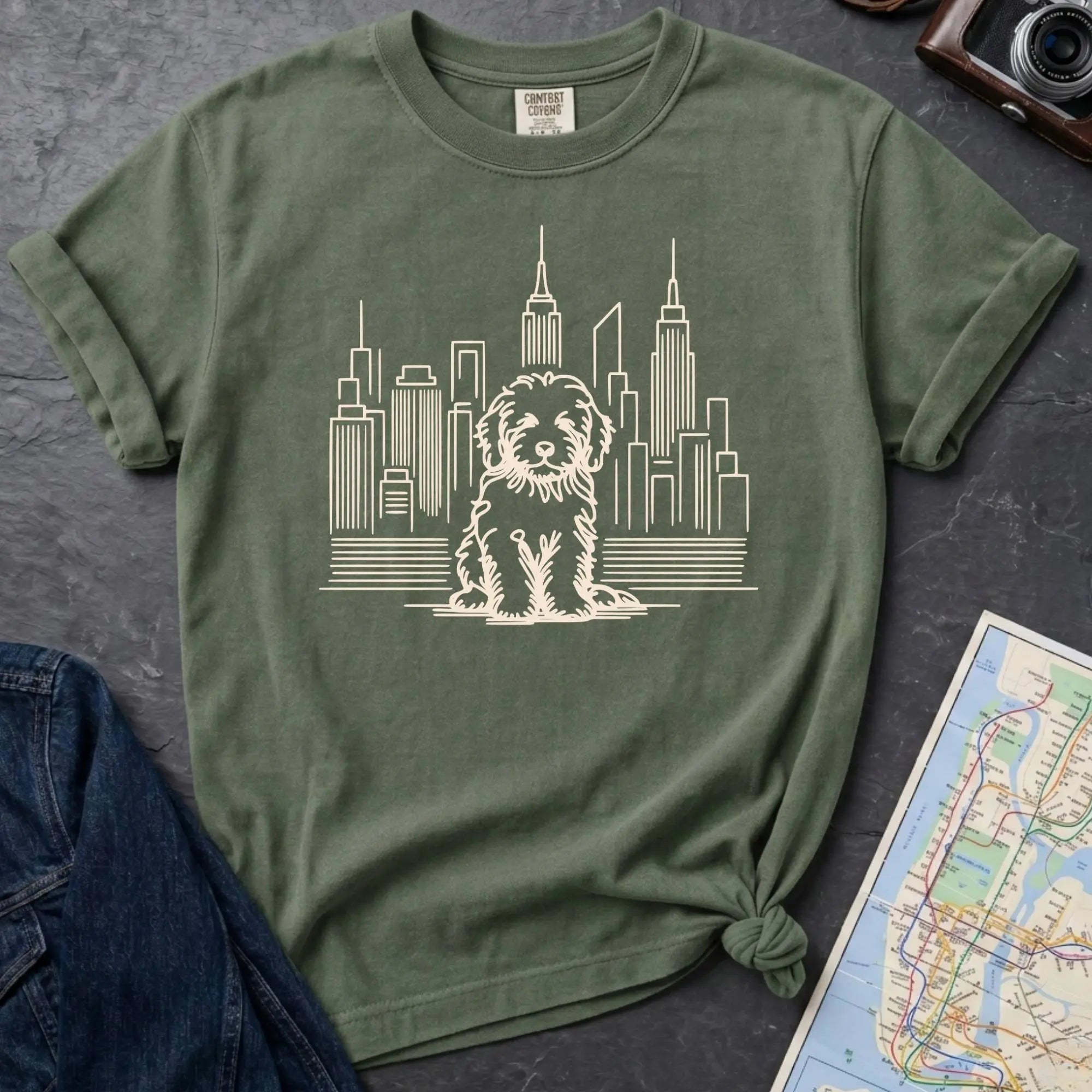Moss green t-shirt with a Golden Doodle and cityscape design on a dark surface with a map and camera.