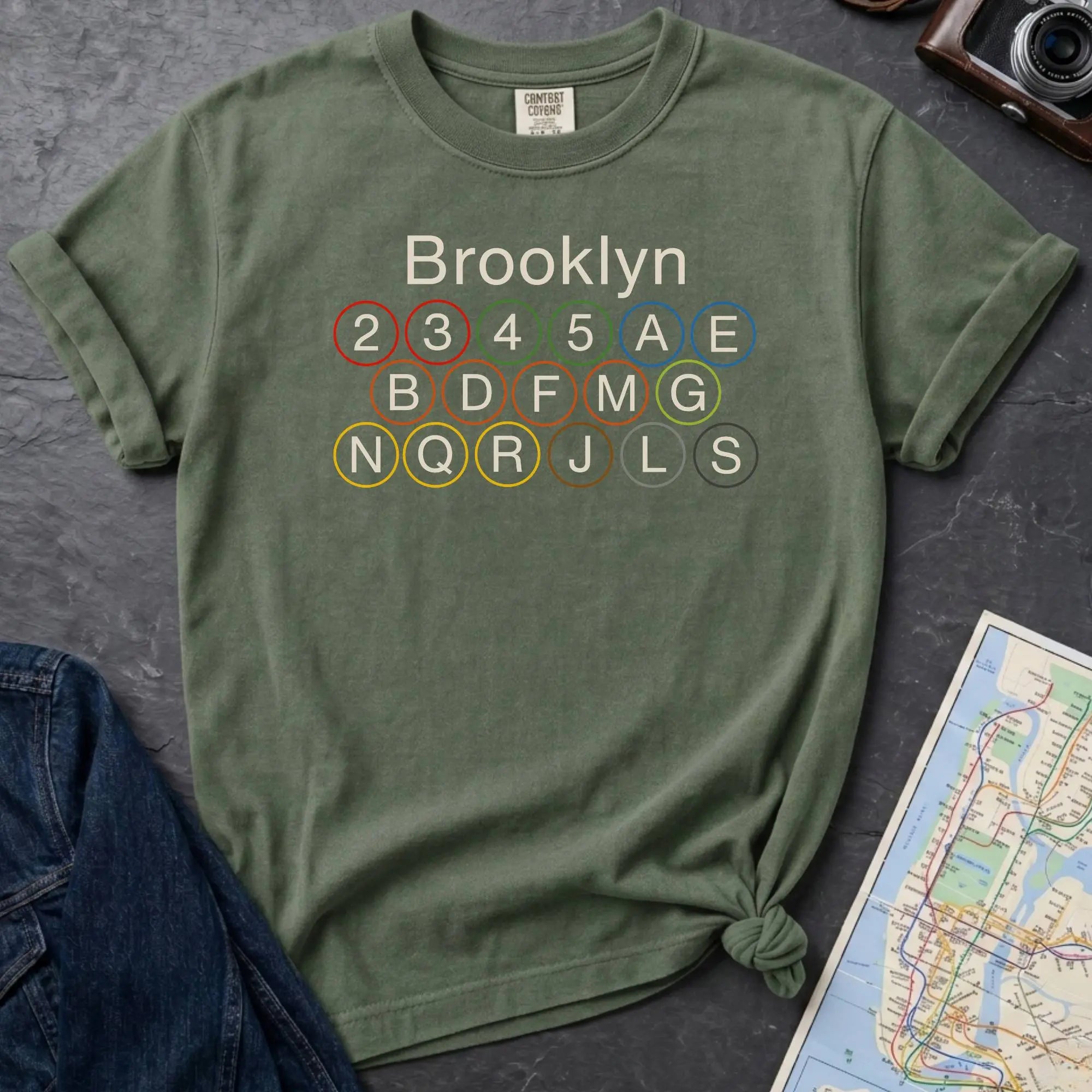 Moss green Comfort Colors t-shirt featuring the word “Brooklyn” above colorful NYC subway line circles representing the subway routes that run through Brooklyn.