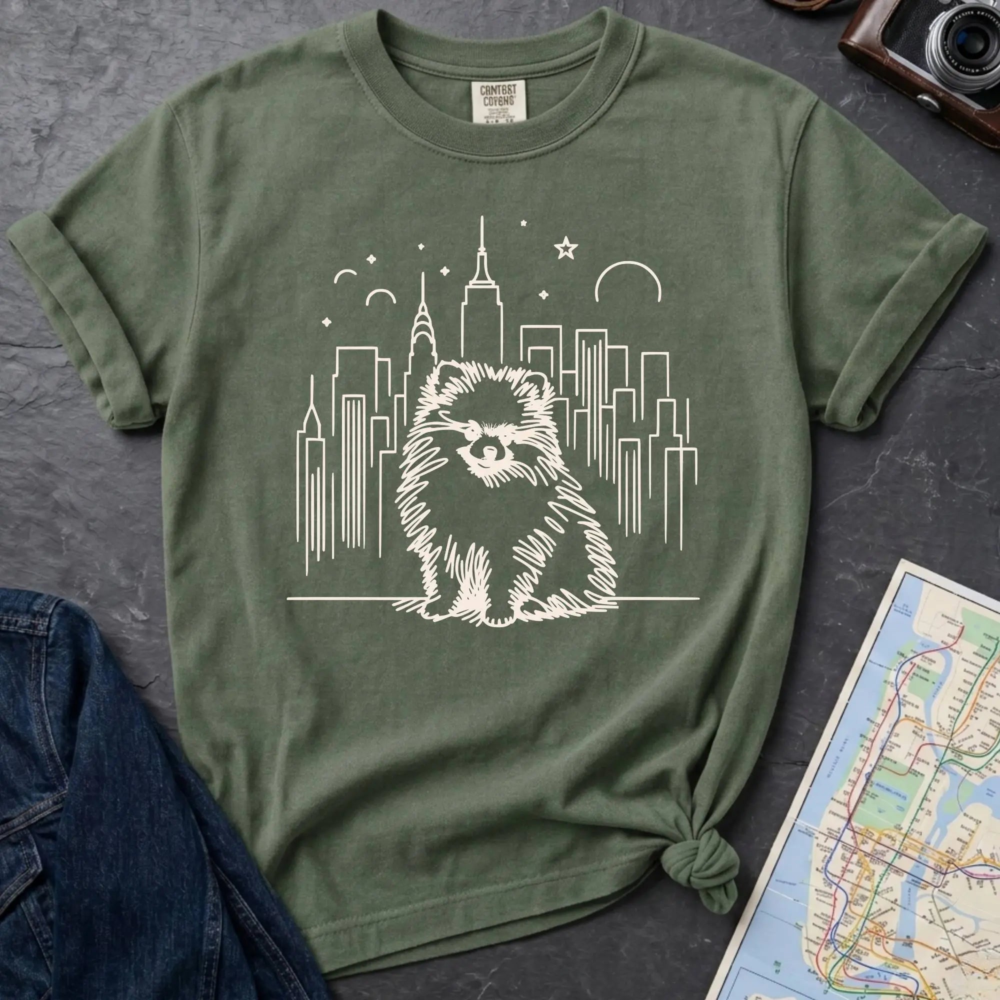 Moss green Comfort Colors t-shirt featuring a fluffy small dog illustration in front of the New York City skyline.