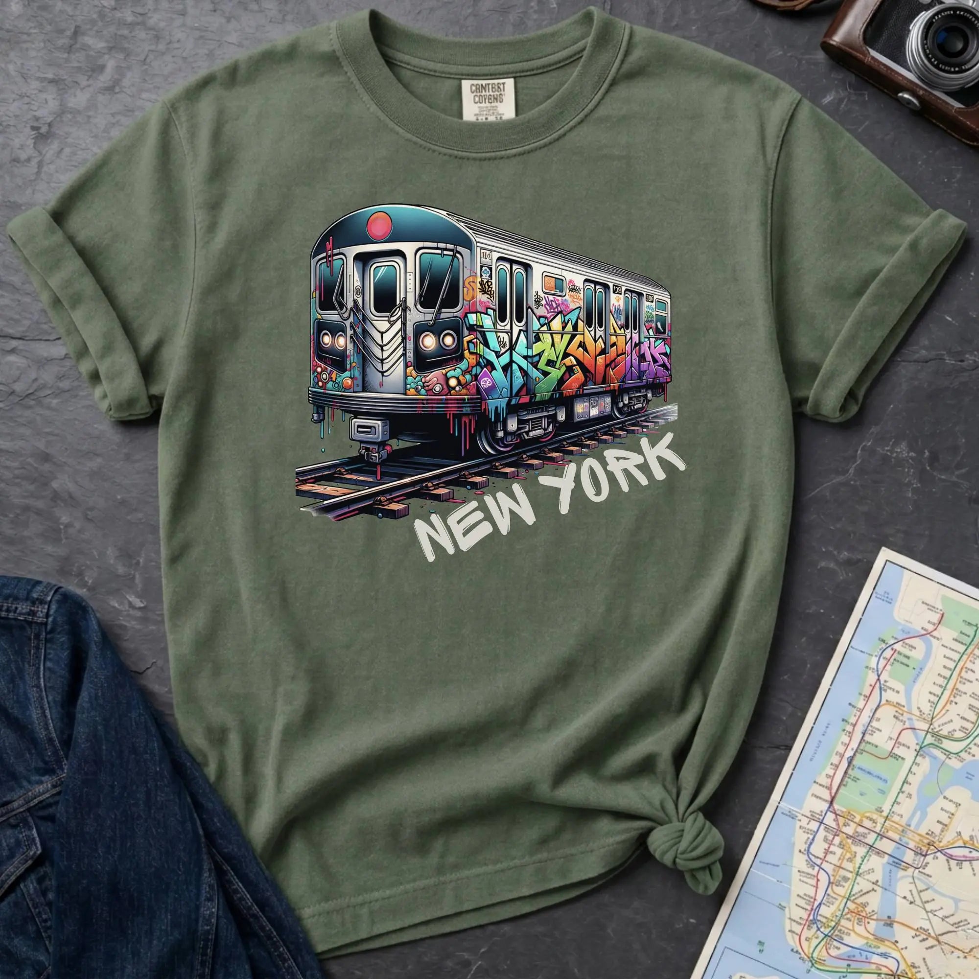 Moss green t-shirt with a colorful train graphic and 'New York' text on a gray surface with a map and denim jacket.