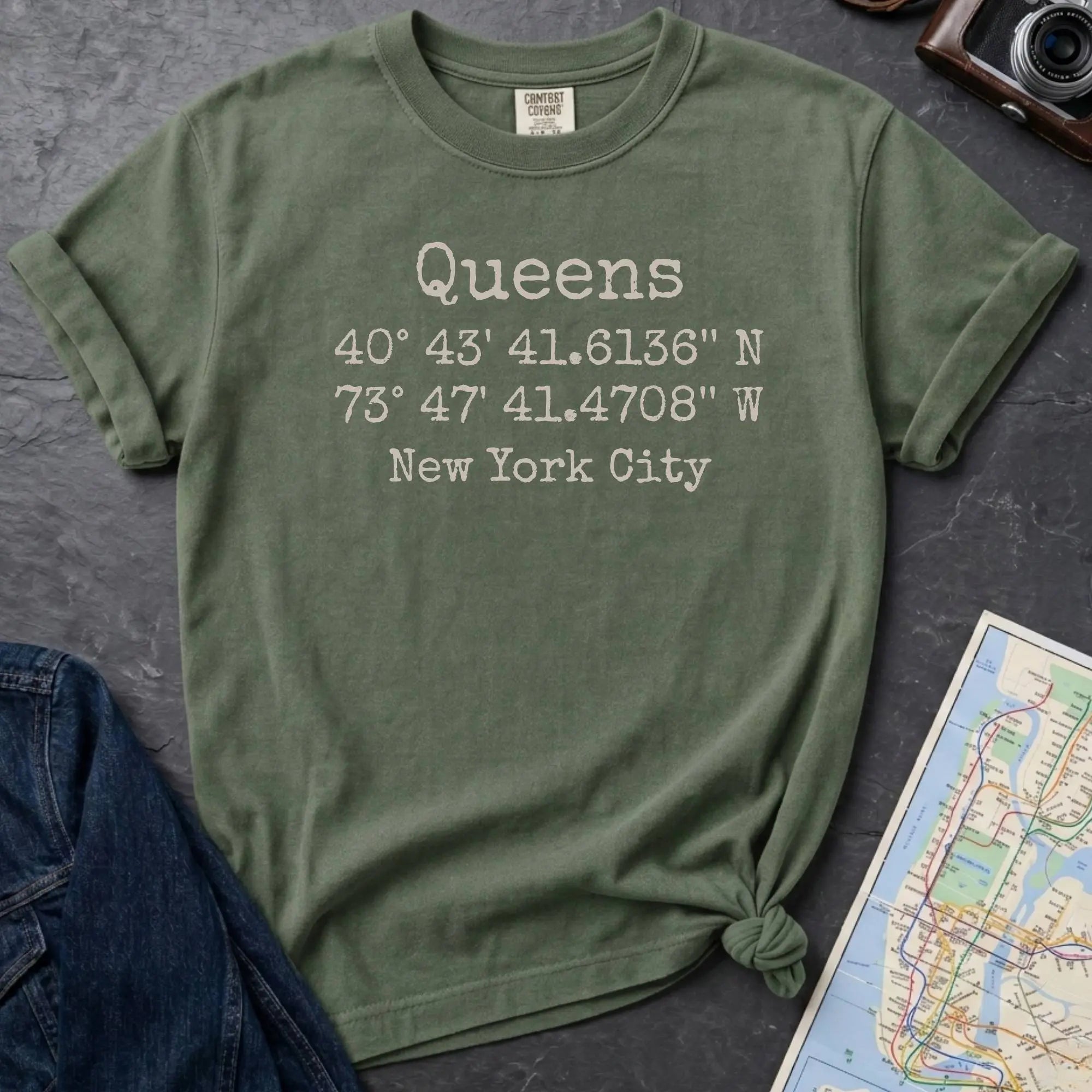 Moss green Comfort Colors t-shirt featuring Queens, NY coordinates and New York City text in a minimalist typewriter-style design.