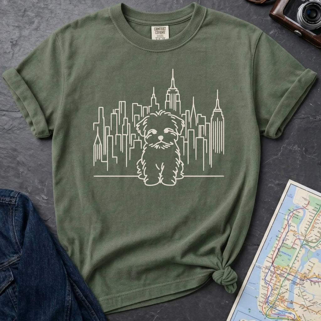 Moss Green t-shirt with a Maltese dog and cityscape design on a gray surface with a map and camera.