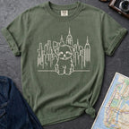 Moss Green t-shirt with a Maltese dog and cityscape design on a gray surface with a map and camera.