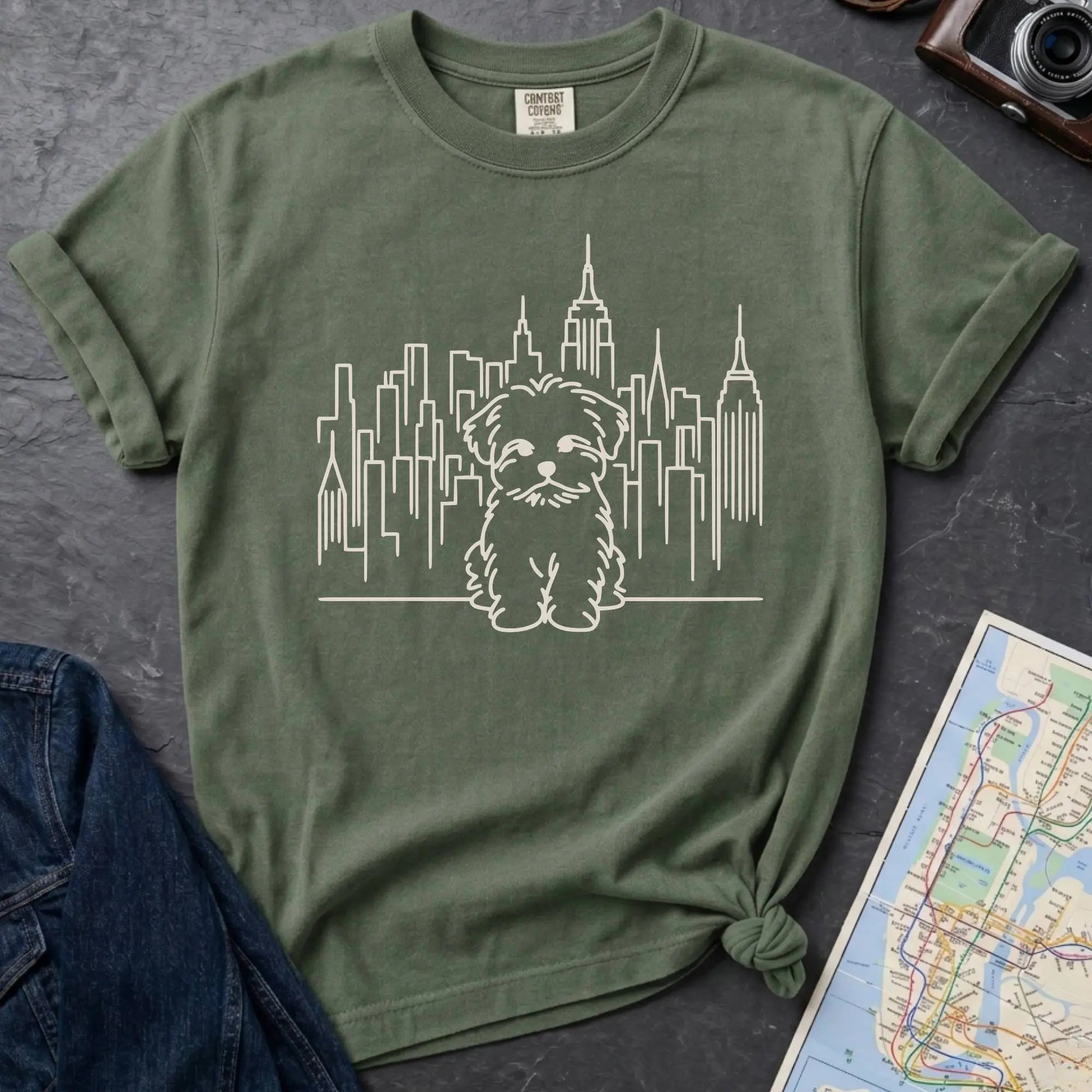 Moss Green t-shirt with a Maltese dog and cityscape design on a gray surface with a map and camera.