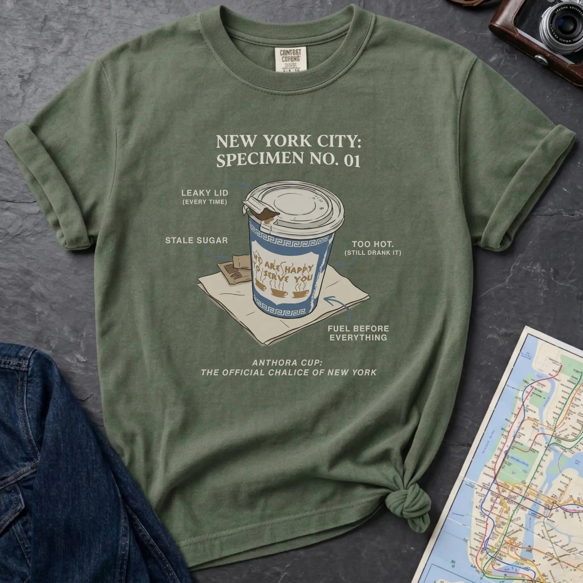 Moss Green t-shirt with New York City coffee cup graphic on a textured surface with map and camera.